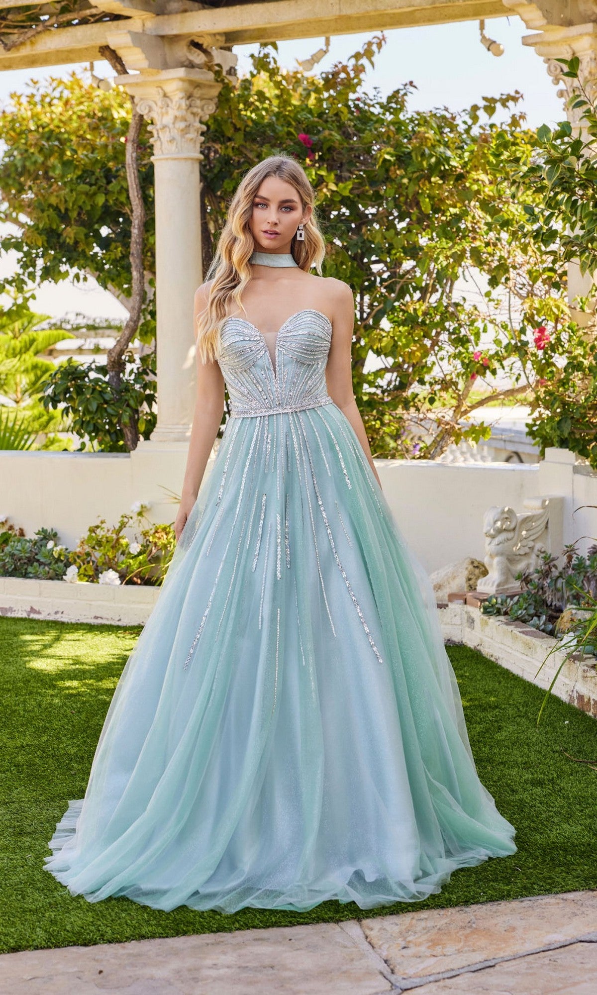 A woman stands outdoors in the Juliet JT2620U long prom dress—light blue, strapless, a-line with a rhinestone-embellished bodice and sparkly accents. Lush greenery and stone columns form the backdrop.